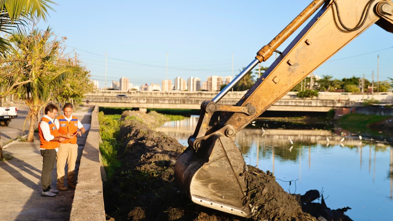 prefeito e coordenador de defesa civil ao lado de canal onde há uma máquina retirando lixo
